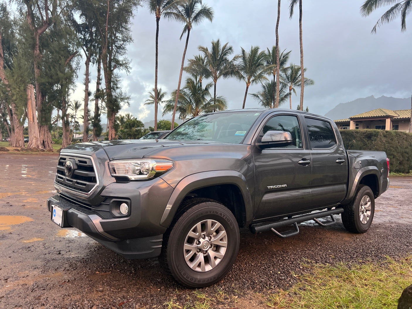 A dark grey Toyota Tacoma pickup truck on a gravel lot after rain, surrounded by tropical trees. Perfect for trips, or consider an rv rental.