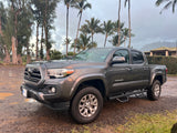 A dark grey Toyota Tacoma pickup truck on a gravel lot after rain, surrounded by tropical trees. Perfect for trips, or consider an rv rental.