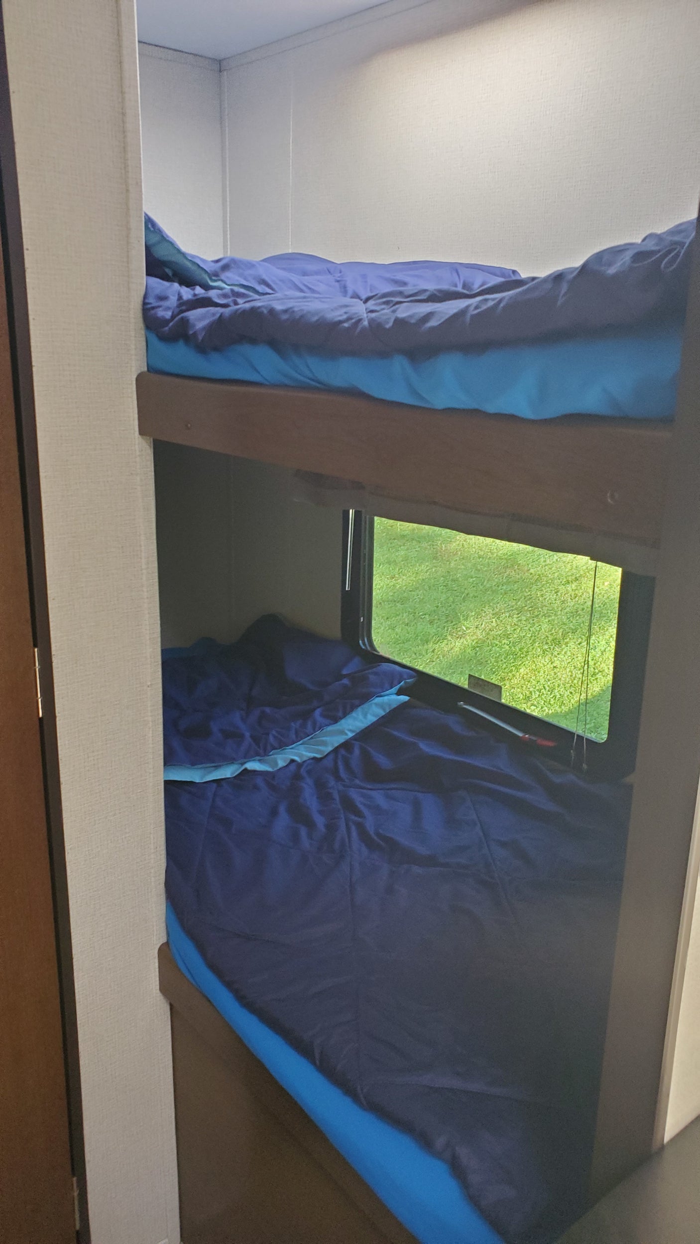 Inside an RV rental: two compact bunk beds with blue bedding. Lower bed features a window overlooking green grass.
