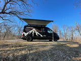 This black **rv rental** camper van, awning out, sits in a dry wooded area under a clear blue sky.