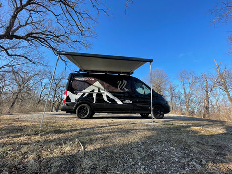 This black **rv rental** camper van, awning out, sits in a dry wooded area under a clear blue sky.