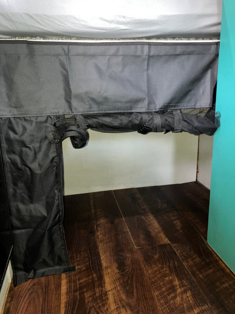 RV rental interior: Black fabric curtain with mesh sections & adjustable straps, secured under a white bed for under-bed storage or pet barrier, on a wooden floor.