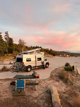 An RV rental camper van with bikes at a mountain campsite, under a stunning pink and blue sunset sky.