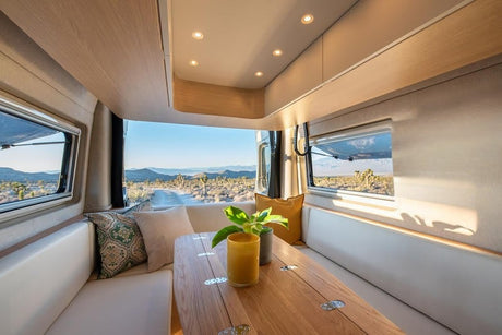 Cozy RV rental interior with light wood, white seating, and wide windows revealing a sunny desert road trip view.