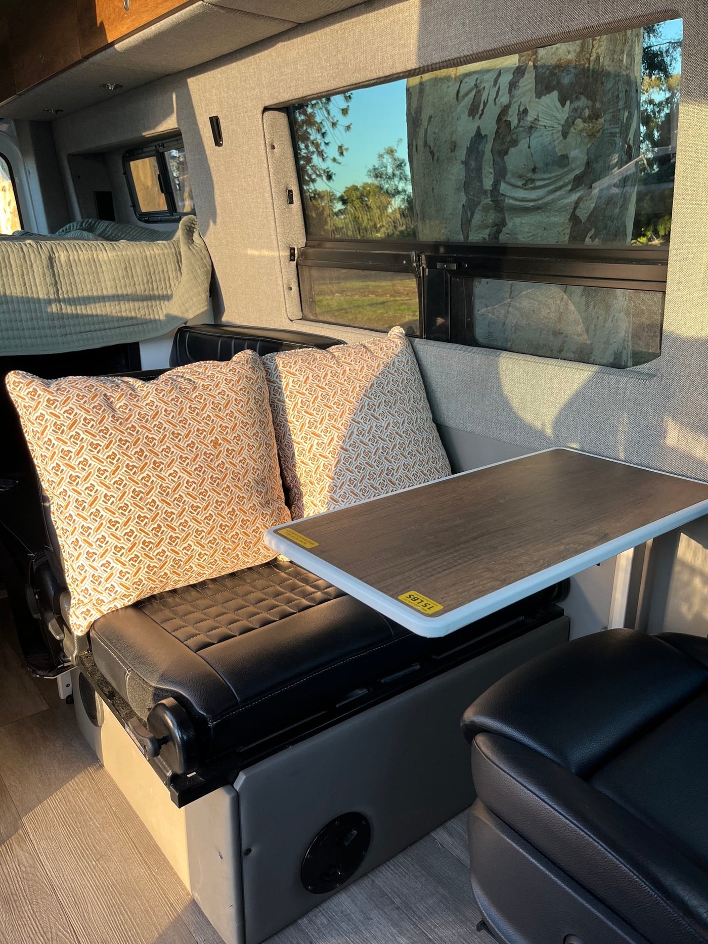 Cozy RV rental interior showing a bed, patterned pillows, table, and seating by a window with tree views. — photo 9