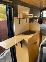Modern RV rental interior: a custom-built van kitchen with wood cabinets, tiled backsplash, sink, and extendable table.
