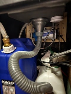 RV rental under-sink plumbing: A flexible hose from the sink drains gray water into a blue tank, with a water pump mounted behind.