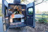 Open rear of a modern RV rental, revealing a bed, utility access, and nature view through a bug screen.