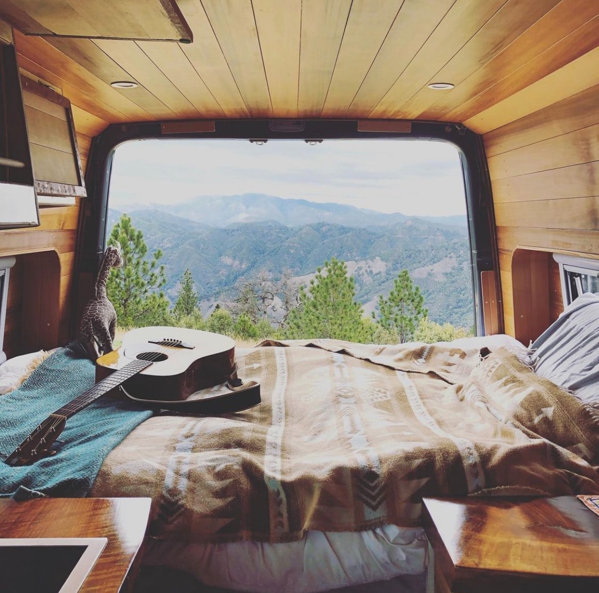 Experience van life: Cozy RV rental bedroom with guitar & giraffe, offering panoramic mountain views from open rear doors.