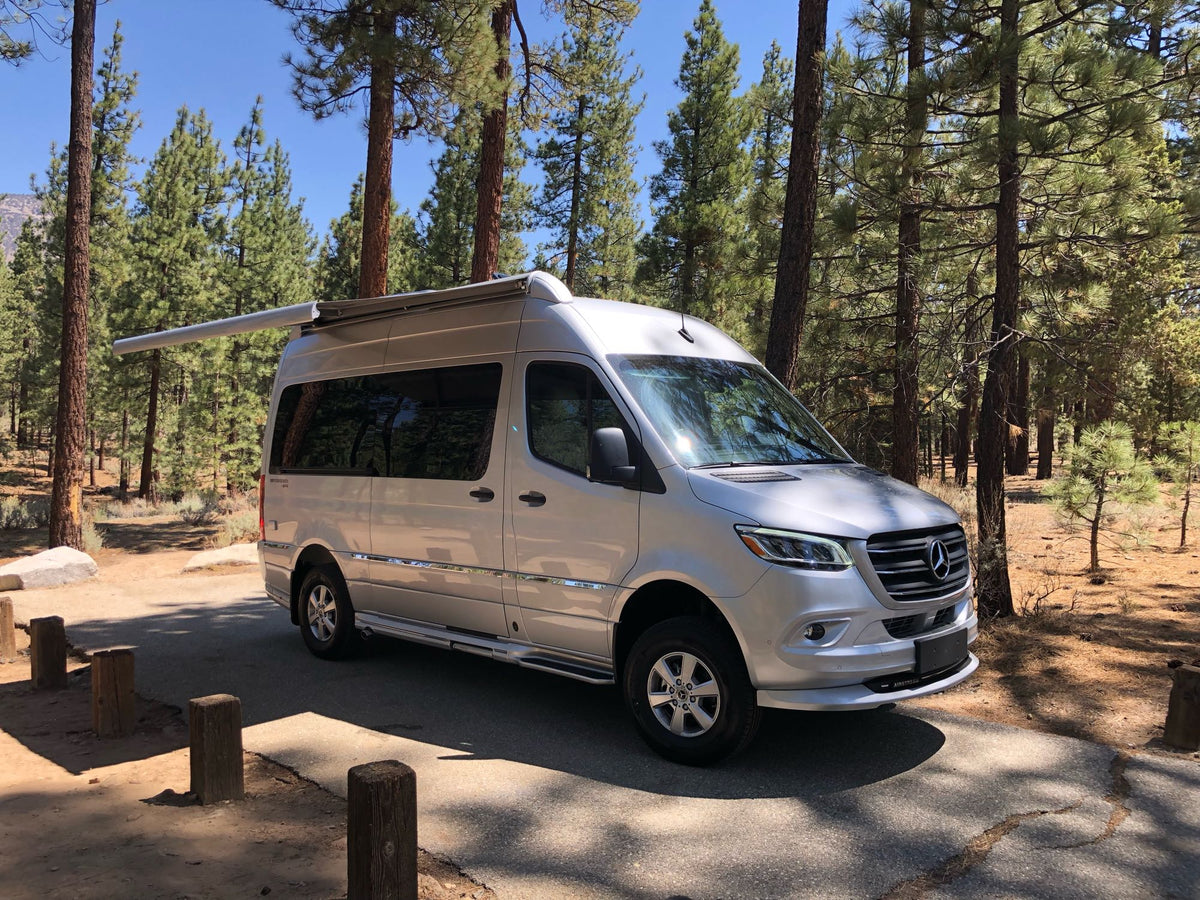 Spacious silver Mercedes-Benz Sprinter van RV rental parked at a serene forest campsite with its awning extended.|Silver Mercedes Sprinter RV rental camper van parked in a sunny pine forest campsite.|Experience outdoor living! This silver rv rental is parked with awning extended and chairs out in a peaceful forest campsite.|Silver Airstream RV with awning extended at a scenic mountain campsite
