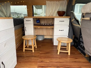 This RV rental shows a camper van's cozy interior, featuring a wooden floor, custom cabinets with two stools, and a bed area for mobile living.