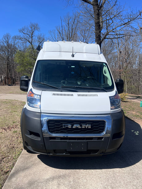 Front view of a white Ram ProMaster van, a perfect RV rental. Parked on concrete in daylight with trees behind.