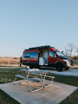 Red RV rental camper van parked at a serene campsite with open doors revealing its interior, a picnic table with coffee nearby.