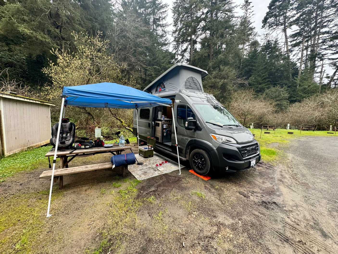 An RV rental camper van with a pop-top tent, blue canopy, and picnic table set up on a muddy campsite.
