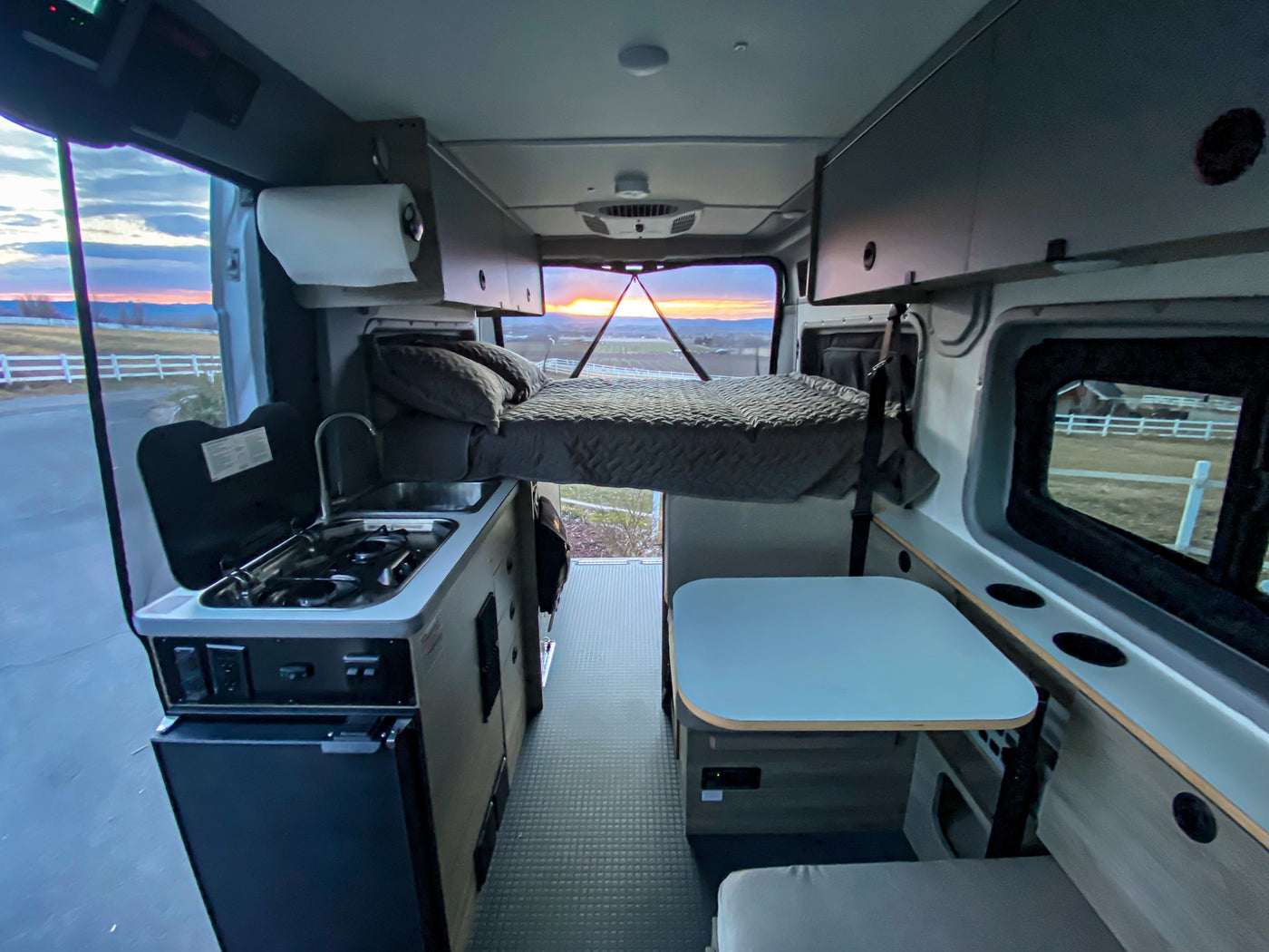 Inside a spacious RV rental: full kitchen, elevated bed, and dining area with a beautiful sunset view from the back.