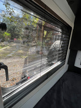View from an RV rental window, with screen and blinds, showing a sunny outdoor scene with trees and a rear-mounted spare tire.