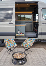 RV rental setup: A camper van with its door open, showing a kitchen, two outdoor chairs, and a fire pit on a rug.