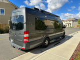 Stylish gray camper van, perfect for an RV rental, parked on a sunny residential street. Features include roof AC and awning.