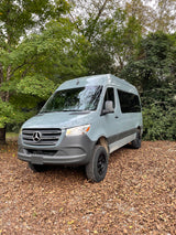 A light grey Mercedes-Benz Sprinter van, ideal for rv rental, is parked on brown autumn leaves amidst green trees.