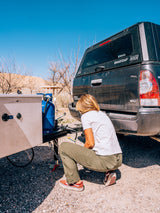 Crouching by an RV rental, a person connects the trailer hitch to a truck, propane tank visible, for outdoor adventure.