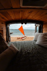 View from an RV rental bed: a serene beach with an orange-sailed boat. Perfect beach camping getaway.