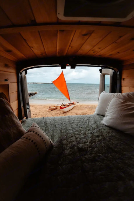 View from an RV rental bed: a serene beach with an orange-sailed boat. Perfect beach camping getaway.