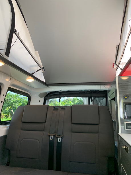**RV rental interior**: Shows a raised pop-top roof, dark grey bench seats with seatbelts, and windows looking out to lush trees.