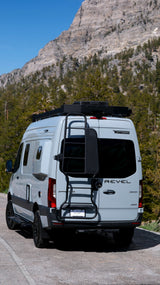 Light gray Winnebago Revel rv rental van parked on a road with rocky mountains and pine trees in the background.