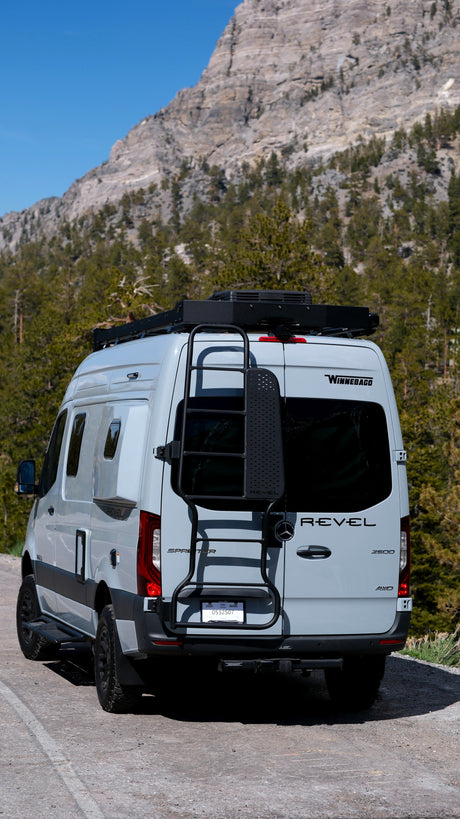 Light gray Winnebago Revel rv rental van parked on a road with rocky mountains and pine trees in the background.