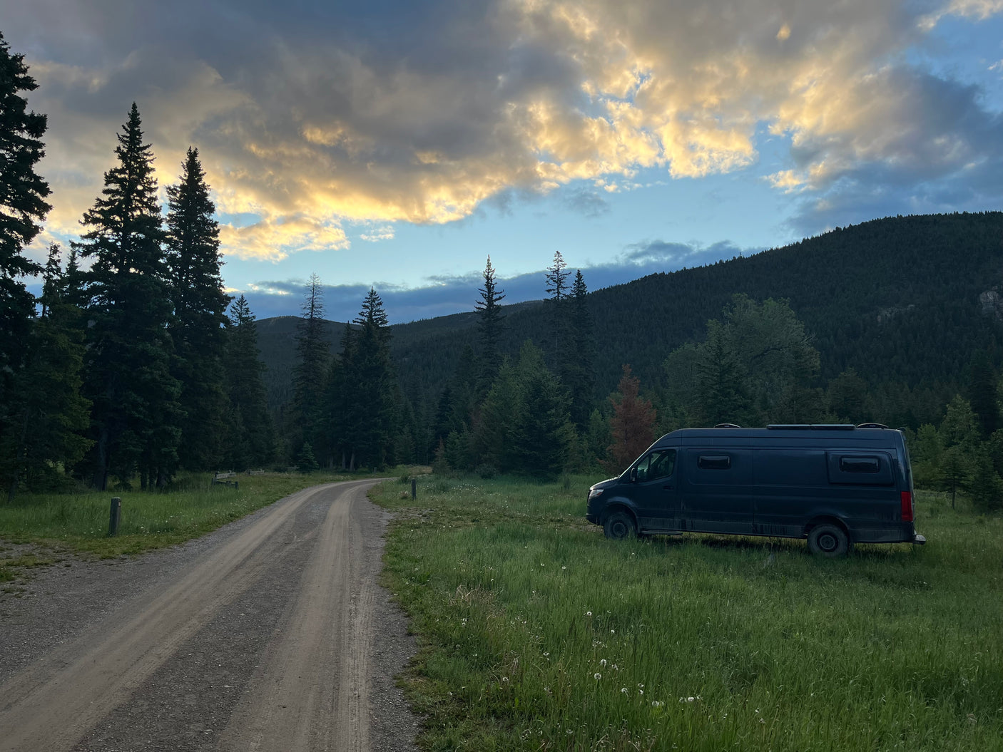 An RV rental camper van in a forest clearing by a dirt road, with trees and mountains under a cloudy, golden sky.