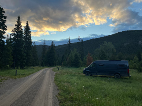 An RV rental camper van in a forest clearing by a dirt road, with trees and mountains under a cloudy, golden sky.