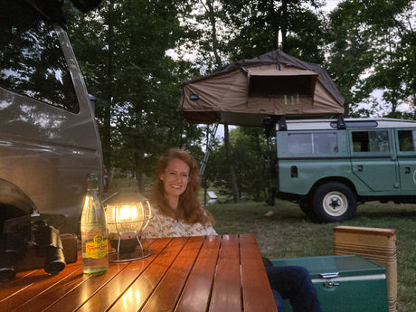 Smiling woman at camp table. Vehicles: a light-colored RV rental van & a green Land Rover with a rooftop tent.