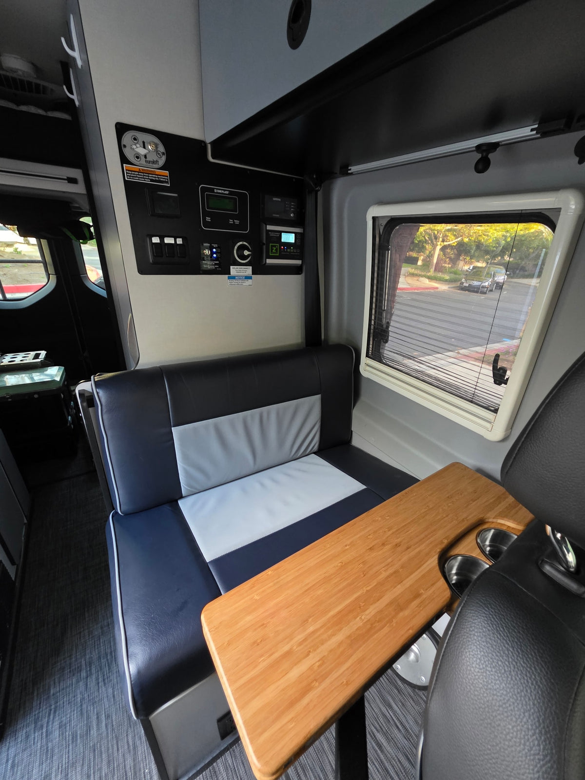 Comfortable camper van rental dinette, featuring a multi-colored bench, wooden table with cup holders, controls, and a window. — photo 8