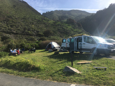 RV rental camper van and tent at a sunny campsite with people, nestled in lush green hills.