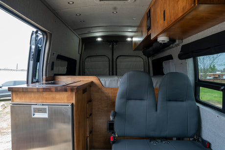 View inside an RV rental: features a wood-top kitchenette, grey seating, bed area, and overhead cabinets.
