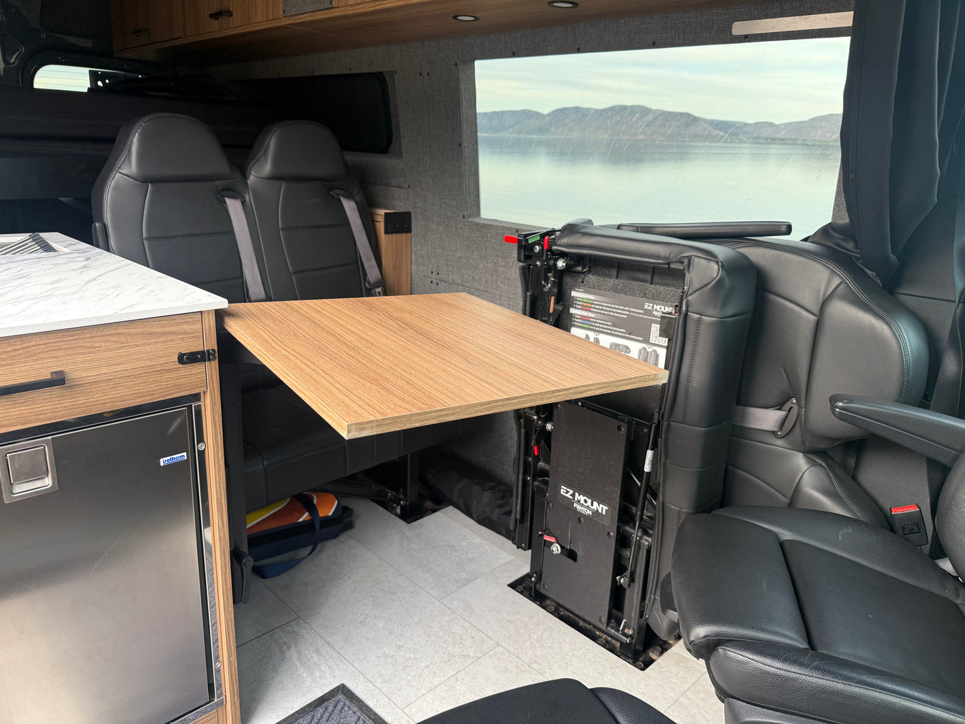 This RV rental interior features comfortable seating, a versatile table, an Isotherm fridge, and a stunning lake view.
