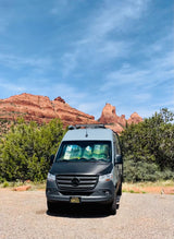 A grey RV rental van, front view, parked on gravel with stunning red rock mountains and green shrubs in the background. Perfect for adventure.