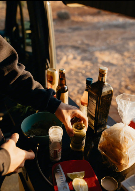Hands prep a camping meal at an RV rental site. Bottles of oil, sauces, salt, and fresh lime on a counter in the desert.