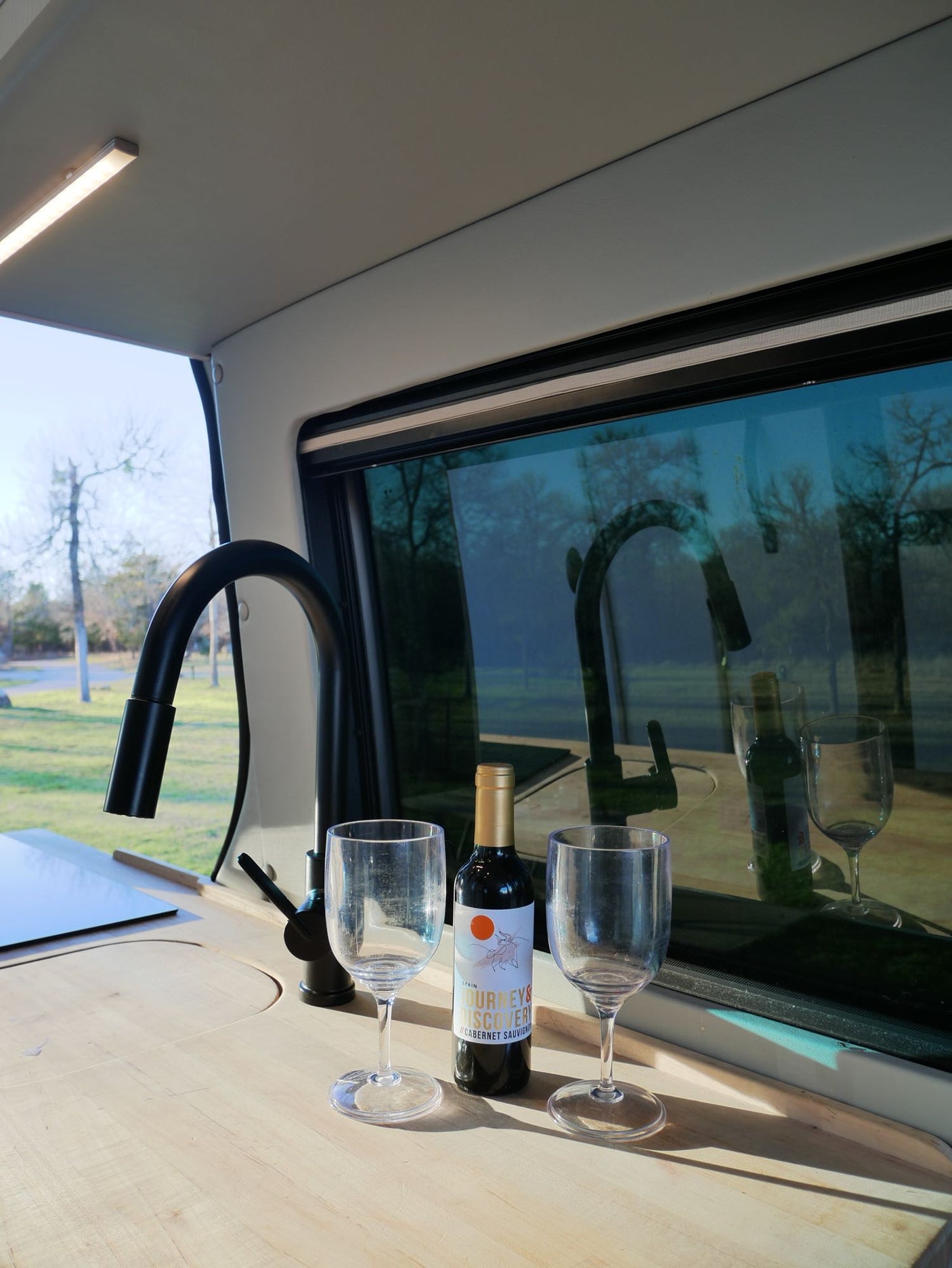 Inside an RV rental: a wooden counter holds wine, two glasses, and a black faucet, reflected in the scenic window overlooking trees.