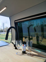 Inside an RV rental: a wooden counter holds wine, two glasses, and a black faucet, reflected in the scenic window overlooking trees.