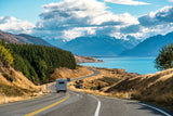 An RV rental drives a winding road beside a turquoise lake, with snow-capped mountains and clouds in the distance. Ideal road trip.
