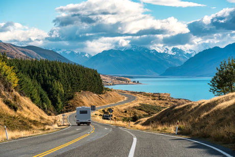 An RV rental drives a winding road beside a turquoise lake, with snow-capped mountains and clouds in the distance. Ideal road trip.