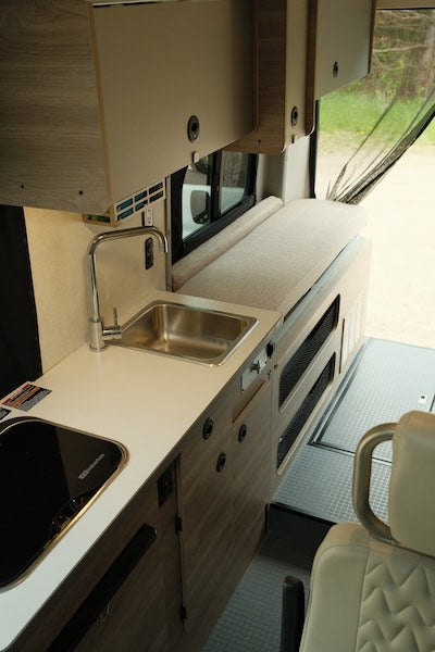 RV rental interior: view of a light-toned kitchenette with sink, induction cooktop, overhead cabinets, and side bench.