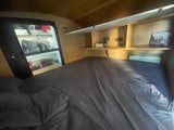 Cozy RV rental interior featuring a bed with sheets, built-in shelves, and an AC unit for comfortable travels.