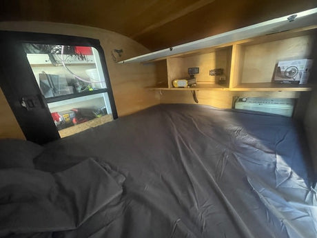 Cozy RV rental interior featuring a bed with sheets, built-in shelves, and an AC unit for comfortable travels.