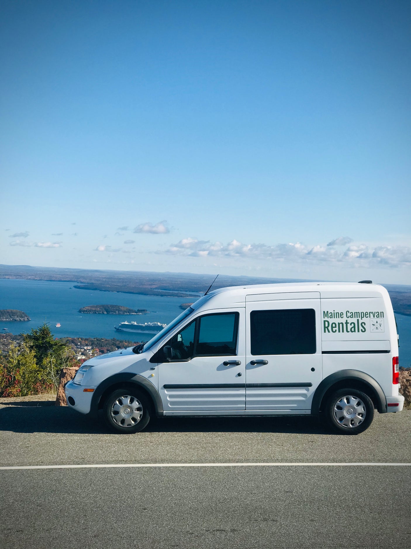 White "Maine Campervan Rentals" van, an ideal rv rental, parked at a scenic overlook with a vast bay, islands, and a cruise ship.