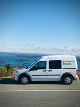 White "Maine Campervan Rentals" van, an ideal rv rental, parked at a scenic overlook with a vast bay, islands, and a cruise ship.