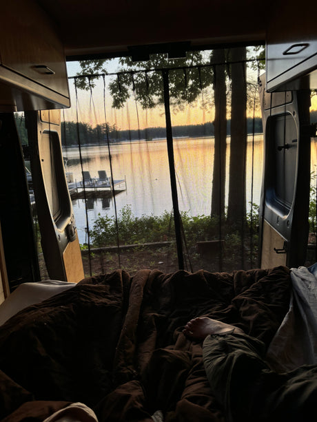 Peaceful rv rental experience: feet propped on a cozy bed, gazing at a stunning lake sunset through a bug screen.