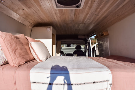 Inside a cozy RV rental van, showcasing a bed with pink bedding, wood-paneled ceiling, and a bright front cab view.