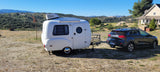 This compact RV rental, towed by a Kia Niro, offers a cozy outdoor escape amidst scenic hills & a distant house.